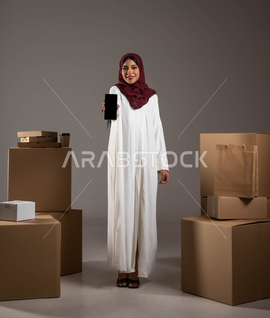 أداء العمل بدقة وإتقان، طلب المنتجات عبر تطبيقات الهاتف المحمول، بورتريه لامرأة عربية خليجية سعودية ترتدي عباءة بيضاء وحجاب أحمر، عرض شاشة سوداء فارغة عبر الموبايل، الشراء والتسوق الأونلاين، خدمات الشحن ذات الجودة العالية، الطول الكامل للجسم، خلفية رمادية