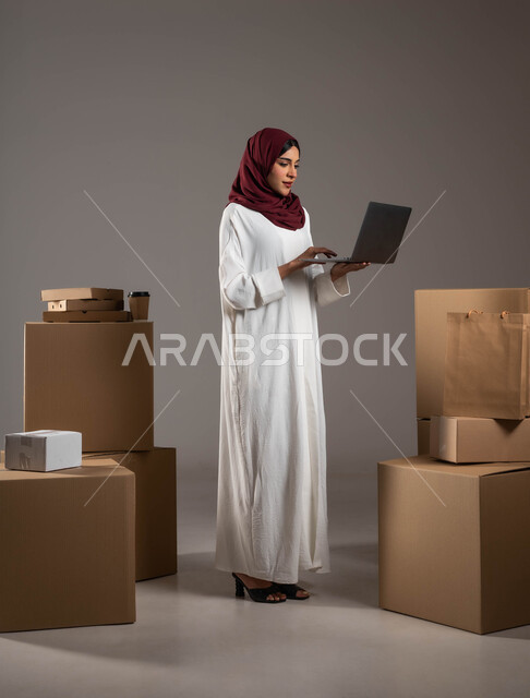 الاهتمام بآراء العملاء، استخدام التقنيات الحديثة في فرز المنتجات وتصنيفها، التأكد من وجود البضائع وتوافرها، بورتريه لامرأة عربية خليجية سعودية ترتدي عباءة بيضاء وحجاب أحمر تعمل في مستودع لتوصيل البضاعة، شحن وتوصيل الطلبات، الطول الكامل للجسم، خلفية رمادية