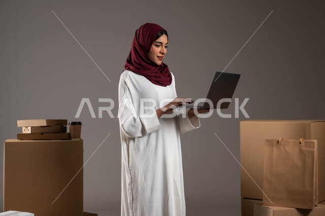 Using modern technologies in sorting and classifying products, ensuring the presence and availability of goods, a portrait of a Saudi Arabian Gulf woman wearing a white abaya and a red veil working in a warehouse to deliver goods, shipping and delivering online orders, paying attention to customer opinions, gray background