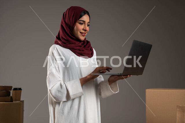 Ensuring the presence and availability of goods, a close-up portrait of a Saudi Gulf Arab woman wearing a white abaya and red veil working in a warehouse to deliver goods, using modern technologies in sorting and classifying products, shipping and delivering online orders, paying attention to customer opinions, gray background