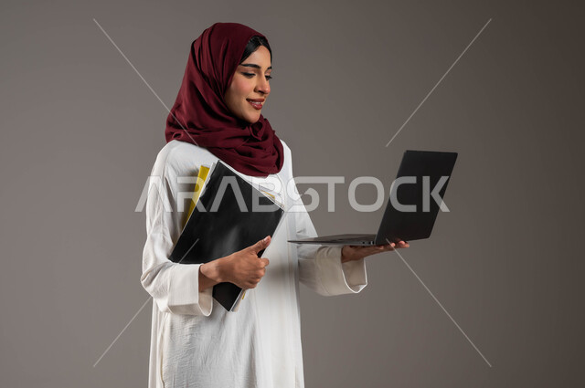 بحث وايجاد فكرة ما عبر الانترنت، استخدام الأجهزة والتقنيات الحديثة في التعلم عن بعد، بورتريه مقرب لشابة عربية خليجية سعودية ترتدي عباءة بيضاء وحجاب أحمر تحمل بيدها ملفات المحاضرات وتقوم بتصفح الحاسوب المحمول، متابعة الحصص الدراسية أونلاين، خلفية رمادية