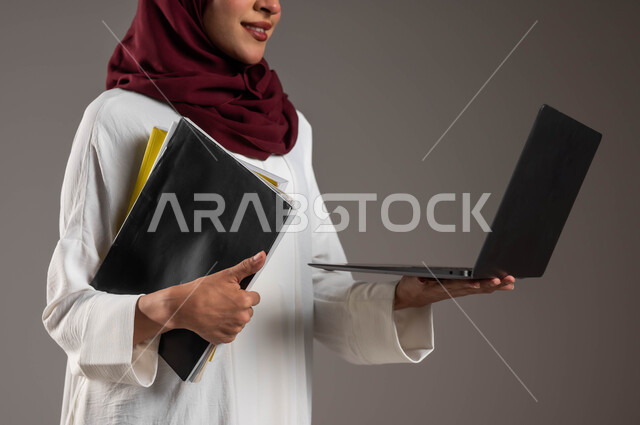 Using modern devices and technologies in distance learning, researching and finding an idea via the Internet, a close-up portrait of a young Saudi Gulf Arab woman wearing a white abaya and a red veil, holding lecture files in her hand and browsing the laptop, following the lessons online, gray background.