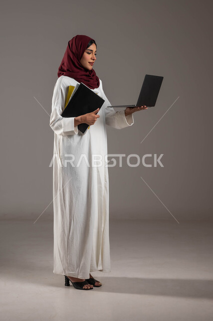 Creating an idea via the Internet, using modern technology devices in distance learning, a portrait of a Saudi Gulf Arab young woman wearing a white abaya and a red hijab, holding lecture files in her hand and browsing the laptop, following classes online, full length of the body, gray background