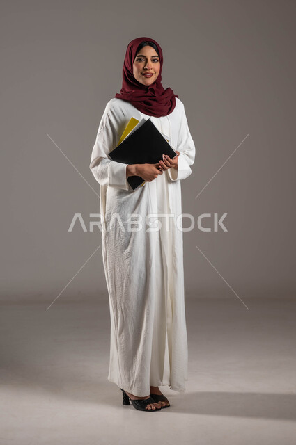 الاستعداد للعام الدراسي الجامعي الجديد، مفهوم التعليم الأكاديمي في الجامعات السعودية، بورتريه لطالبة جامعية عربية خليجية سعودية ترتدي عباءة بيضاء وحجاب أحمر تحمل بيدها ملفات المحاضرات، النظر الى الكاميرا بإيماءات الفرح والسرور، الطول الكامل للجسم، خلفية رمادية