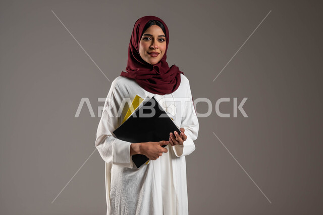 مفهوم التعليم الأكاديمي في الجامعات السعودية، الاستعداد للعام الدراسي الجامعي الجديد، بورتريه لطالبة جامعية عربية خليجية سعودية ترتدي عباءة بيضاء وحجاب أحمر تحمل بيدها كتب وملفات المحاضرات، النظر الى الكاميرا بإيماءات الفرح والسرور، خلفية رمادية
