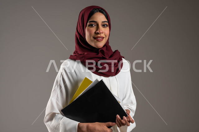النظر الى الكاميرا بإيماءات الفرح والسرور، الاستعداد للعام الدراسي الجامعي الجديد، مفهوم التعليم الأكاديمي في الجامعات السعودية، بورتريه مقرب لطالبة جامعية عربية خليجية سعودية ترتدي عباءة بيضاء وحجاب أحمر تحمل بيدها ملفات المحاضرات، خلفية رمادية