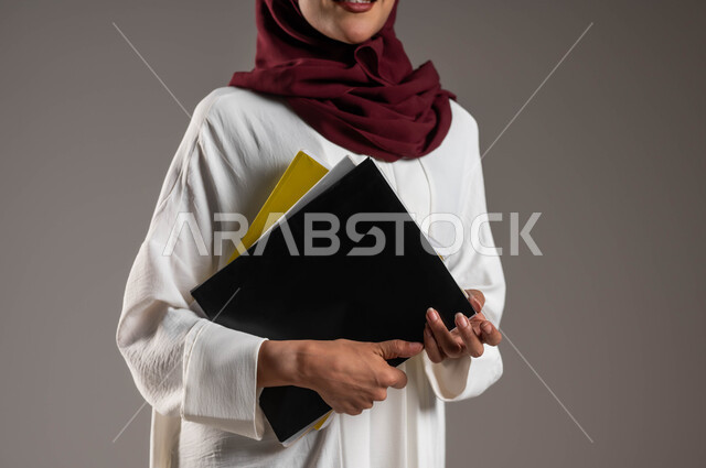 التعلم والتعليم الأكاديمي في الجامعات السعودية، الاستعداد للعام الدراسي الجامعي الجديد، بورتريه مقرب لطالبة جامعية عربية خليجية سعودية ترتدي عباءة بيضاء وحجاب أحمر تحمل بيدها ملفات المحاضرات، خلفية رمادية