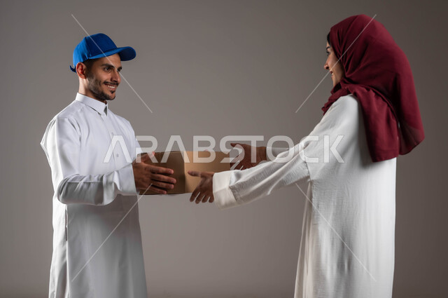 Ordering purchases online, the process of transferring orders to customers, smart and safe shopping, a close-up portrait of a Saudi Arabian Gulf woman wearing a white abaya and red hijab receiving a cardboard box, a delivery person delivering products, gray background