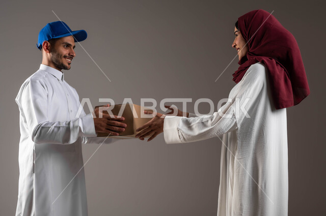 Reliable delivery services, ordering purchases online, safe smart shopping, a close-up portrait of a Saudi Arabian Gulf woman wearing a white abaya and red hijab receiving a cardboard box, a sales representative delivering products, gray background