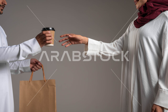 التسوق الذكي عبر الاونلاين، ضمان تسليم المشروبات الساخنة، بورتريه مقرب لامرأة عربية خليجية سعودية ترتدي عباءة بيضاء وحجاب أحمر تقوم بإستلام القهوة الساخنة وكيس مشتريات كرتون، مندوب مبيعات يقوم بتوصيل المنتجات الى المنزل، التوصيل السريع والإمن، خلفية رمادية