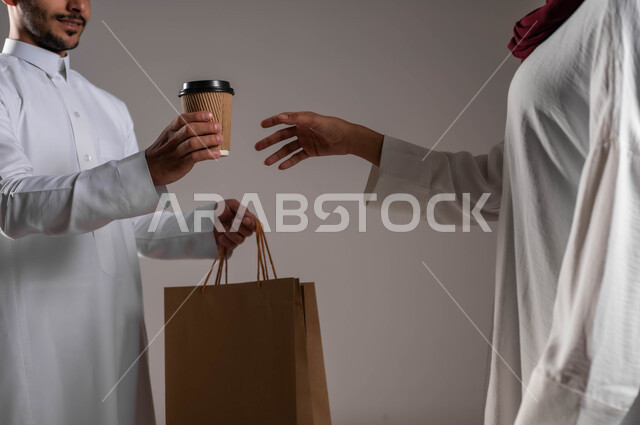 التوصيل السريع والإمن، التسوق الذكي عبر الاونلاين، ضمان تسليم المشروبات الساخنة، بورتريه مقرب لامرأة عربية خليجية سعودية ترتدي عباءة بيضاء وحجاب أحمر تقوم بإستلام القهوة الساخنة وكيس مشتريات كرتون، مندوب مبيعات يقوم بتوصيل المنتجات الى المنزل، خلفية رمادية