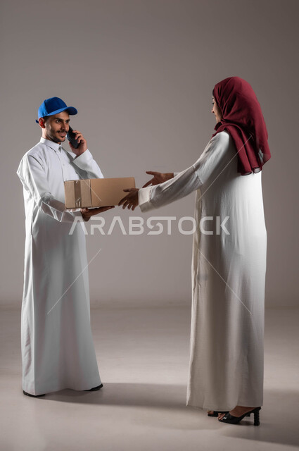 خدمات الشحن والتوصيل السريع والموثوق، ميزة التسوق الالكتروني والطلب أونلاين، بورتريه لامرأة عربية خليجية سعودية ترتدي عباءة بيضاء وحجاب أحمر تستلم صندوق كرتون، مندوب توصيل يقوم بإجراء مكالمة هاتفية للتأكد من المنتجات، الطول الكامل للجسم، خلفية رمادية