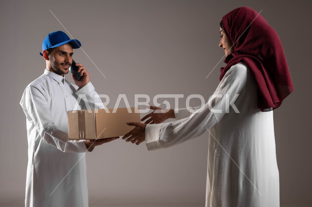 ميزات التسوق الالكتروني والطلب أونلاين، خدمات الشحن والتوصيل السريع والموثوق، بورتريه لامرأة عربية خليجية سعودية ترتدي عباءة بيضاء وحجاب أحمر تستلم صندوق كرتون، مندوب توصيل يقوم بإجراء مكالمة هاتفية للتأكد من المنتجات، خلفية رمادية