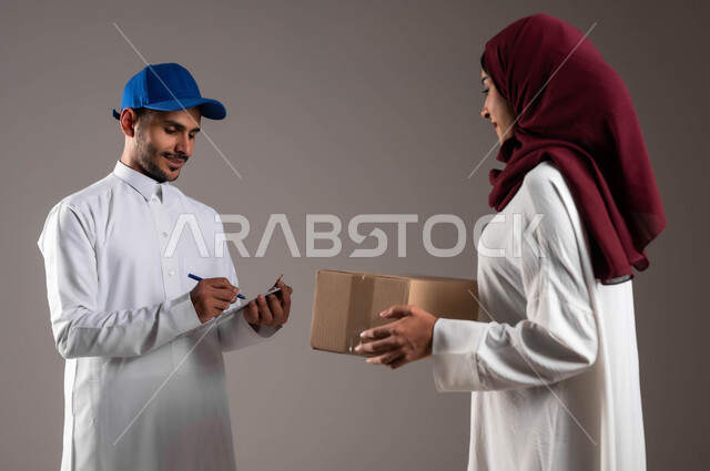 مفهوم التسوق وشراء الحاجيات عبر الاترنت، بورتريه من الجانب لامرأة عربية خليجية سعودية تقوم باستلام الطرد من المندوب، التوقيع على أوراق ومستندات استلام وتسليم الطرود، ضمان سلامة البضائع المرسلة، خدمات التوصيل السريعة في السعودية، خلفية رمادية