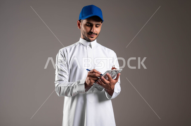 خدمات التوصيل السريعة في السعودية، ضمان سلامة البضائع المرسلة، مفهوم التسوق وشراء الحاجيات عبر الاترنت، بورتريه لشاب عربي خليجي سعودي يرتدي الثوب التقليدي وقبعة زرقاء يقوم باداء عمله، التوقيع على أوراق ومستندات استلام وتسليم الطرود، خلفية رمادية