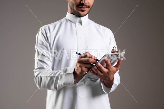 التوقيع على أوراق ومستندات استلام وتسليم الطرود، خدمات التوصيل السريعة في السعودية، ضمان سلامة البضائع المرسلة، مفهوم التسوق وشراء الحاجيات عبر الاترنت، بورتريه مقرب ليد شاب عربي خليجي سعودي يرتدي الثوب التقليدي يقوم باداء عمله، خلفية رمادية