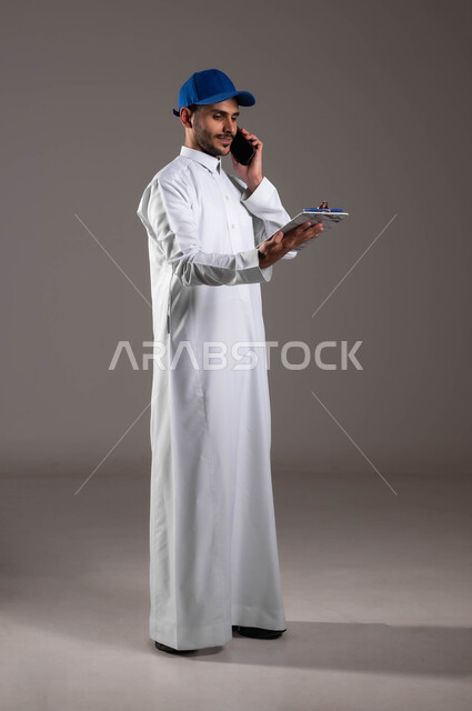 Using modern technology to facilitate work, communicating with customers via mobile phone, close-up portrait from the side of a smiling Saudi Gulf Arab young man wearing a traditional dress and a blue hat looking at papers and documents for receiving and delivering the package, full-body portrait, gray background