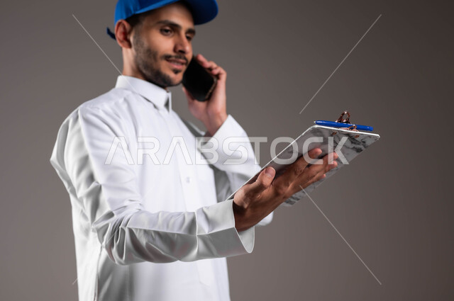 التواصل مع الزبائن عبر الهاتف المحمول، استخدام التكنولوجيا الحديثة، بورتريه مقرب من الجانب لشاب عربي خليجي سعودي مبتسم يرتدي الثوب التقليدي وقبعة زرقاء ينظرإلى أوراق ومستندات استلام وتسليم الطرد، خدمات التوصيل السريعة في السعودية، خلفية رمادية