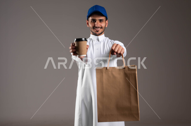 أداء العمل بدقة واتقان، ايماءات السرور والسعادة، توصيل المشروبات الساخنة، تسليم الطلبات والبضائع بأمان وسرعة، مفهوم التسوق عبر الاترنت، بورتريه لشاب عربي خليجي سعودي  يرتدي الزي التقليدي و قبعة زرقاء يحمل بيده أكياس ورقية وكأس، خلفيه رماديه