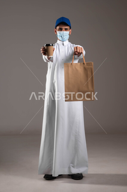 توصيل المشروبات الساخنة، ارتداء الكمامة للوقاية من فايروس كورونا، تسليم الطلبات والبضائع بأمان وسرعة، الالتزام بالجراءات الاحترازية، بورتريه لشاب عربي خليجي سعودي  يرتدي الزي التقليدي و قبعة زرقاء يحمل بيده أكياس تسوق ورقية وكأس، صورة لكامل الجسم، خلفيه رماديه
