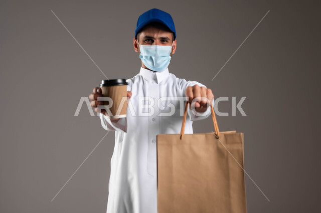 ارتداء الكمامة للوقاية من فايروس كورونا، تسليم الطلبات والبضائع بأمان وسرعة، توصيل المشروبات الساخنة، الالتزام بالجراءات الاحترازية، بورتريه مقرب لشاب عربي خليجي سعودي  يرتدي الزي التقليدي و قبعة زرقاء يحمل بيده أكياس تسوق ورقية وكأس، خلفيه رماديه