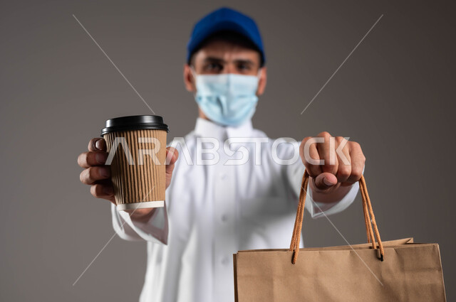 الالتزام بالجراءات الاحترازية، ارتداء الكمامة للوقاية من فايروس كورونا، تسليم الطلبات والبضائع بأمان وسرعة، توصيل المشروبات الساخنة، بورتريه مقرب لشاب عربي خليجي سعودي  يرتدي الزي التقليدي و قبعة زرقاء يحمل بيده أكياس تسوق ورقية وكأس، خلفيه رماديه