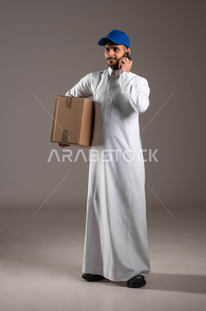 عملية نقل الطلبات، خدمة التوصيل السريعة والآمن في السعودية، استخدام التكنولوجيا الحديثة للمساعدة في العمل، التواصل مع الزبائن عبر الموبايل، بورتريه لشاب عربي خليجي سعودي مبتسم يرتدي الزي التقليدي والقبعة الزرقاء يحمل بيده صندوق، صورة لكامل الجسم، خلفيه رماديه