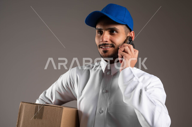 ضمان سلامة الطرود المرسلة، خدمة التوصيل السريعة والآمن في السعودية، استخدام التكنولوجيا الحديثة للمساعدة في العمل، التواصل مع الزبائن عبر الموبايل، بورتريه مقرب لشاب عربي خليجي سعودي مبتسم يرتدي الزي التقليدي والقبعة الزرقاء يحمل بيده صندوق، خلفيه رماديه