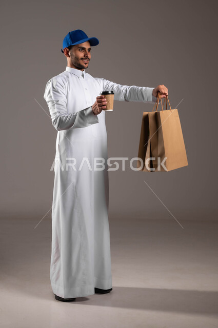 Delivering hot drinks, performing the work with precision and perfection, a portrait of a smiling Saudi Gulf Arab young man wearing a traditional dress and a blue hat, holding paper shopping bags and a cup in his hand, ensuring the safety of sent packages, fast and safe shipping service in Saudi Arabia, a full-body portrait, gray background
