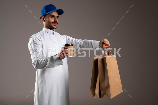 خدمة الشحن السريعة والآمن في السعودية، توصيل المشروبات الساخنة، أداء العمل بدقة واتقان، بورتريه لشاب عربي خليجي سعودي مبتسم يرتدي الزي التقليدي والقبعة الزرقاء يحمل بيده أكياس تسوق ورقية وكأس، ضمان سلامة الطرود المرسلة، خلفيه رماديه