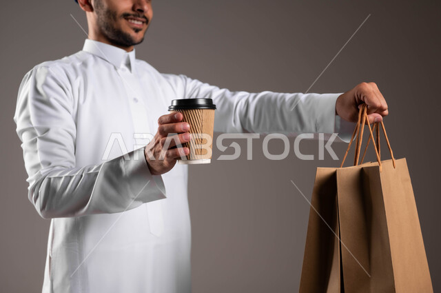 مفهوم التسوق عبر الاترنت، أداء العمل بدقة واتقان، خدمة الشحن السريعة والآمن في السعودية، توصيل المشروبات الساخنة، بورتريه مقرب ليدي شاب عربي خليجي سعودي  يرتدي الزي التقليدي يحمل بيده أكياس تسوق ورقية وكأس، ضمان سلامة الطرود المرسلة، خلفيه رماديه
