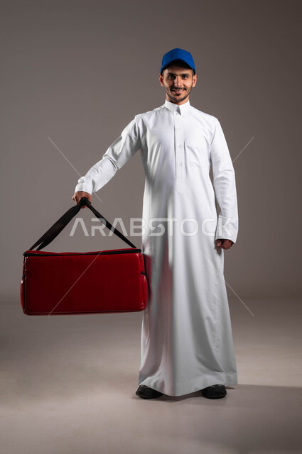 تعابير السرور والسعادة، عملية الشحن والتوصيل الموثوقه، اداء العمل بدقة واتقان، التسوق وشراء الحاجيات عبر الانترنت، ضمان سلامة الطرود المرسلة، بورتريه لشاب عربي خليجي سعودي يرتدي الزي التقليدي وقبعة زرقاء يحمل بيده حقيبة حمراء، صورة لكامل الجسم، خلفية رماديه