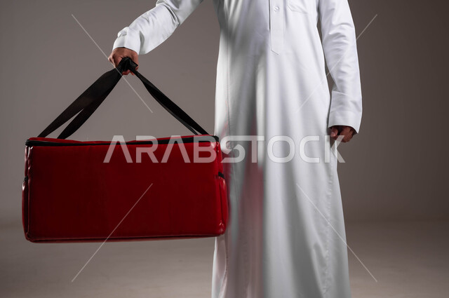 تسليم البضائع للزبائن بسرعة وأمان، عملية الشحن والتوصيل الموثوقه، اداء العمل بدقة واتقان، التسوق وشراء الحاجيات عبر الانترنت، ضمان سلامة الطرود المرسلة، بورتريه مقرب ليد شاب عربي خليجي سعودي يرتدي الزي التقليدي يحمل حقيبة حمراء، خلفية رماديه