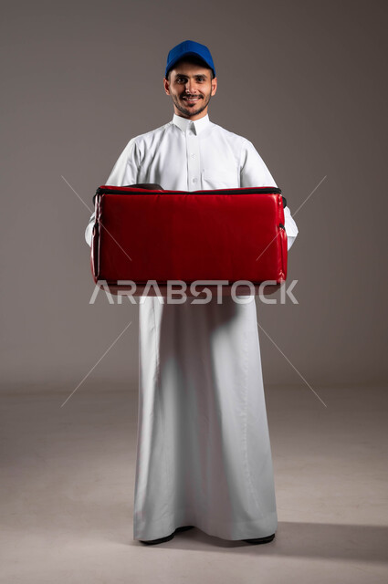 Performing work with precision and mastery, shopping and purchasing necessities online, ensuring the safety of packages and sent goods, portrait of a Saudi Gulf Arab young man wearing a traditional dress and a blue hat holding a red bag in his hand, reliable shipping and delivery process, full body photo, gray background