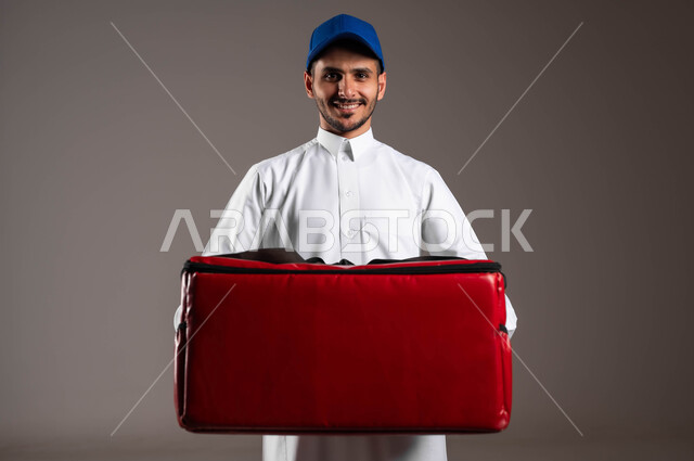 عملية الشحن والتوصيل الموثوقه، اداء العمل بدقة واتقان، التسوق وشراء الحاجيات عبر الانترنت، ضمان سلامة الطرود و البضائع المرسلة، بورتريه مقرب لشاب عربي خليجي سعودي يرتدي الزي التقليدي  وقبعة زرقاء يحمل بيده حقيبة حمراء، خلفية رماديه