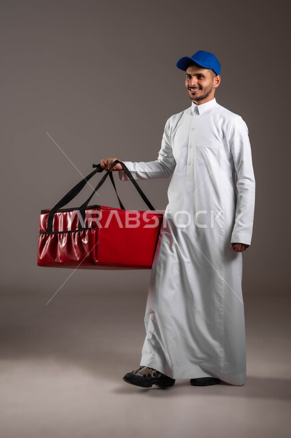 Gestures of happiness and pleasure, ensuring the safety of sent packages and goods, a portrait of a smiling Saudi Gulf Arab young man wearing a traditional dress and a blue hat holding a red bag in his hand, reliable shipping and delivery process, performing work accurately and perfectly, full-length body portrait, gray background