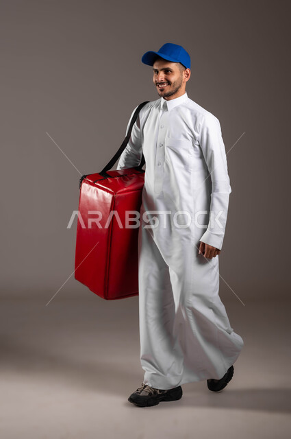 الانطلاق إلى العمل بهمة ونشاط، ضمان سلامة الطرود و البضائع المرسلة، بورتريه  لشاب عربي خليجي سعودي مبتسم يرتدي الزي التقليدي  وقبعة زرقاء يحمل بيده حقيبة حمراء، عملية الشحن والتوصيل الموثوقه، اداء العمل بدقة واتقان، صوره الجسم بالطول الكامل، خلفية رماديه