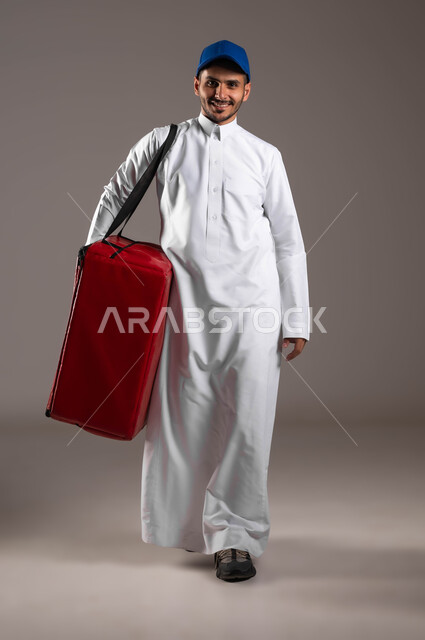 النظر إلى الكاميرا بايماءات السعادة، ضمان سلامة الطرود و البضائع المرسلة، بورتريه  لشاب عربي خليجي سعودي مبتسم يرتدي الزي التقليدي  وقبعة زرقاء يحمل بيده حقيبة حمراء، عملية الشحن والتوصيل الموثوقه، اداء العمل بدقة واتقان، صوره الجسم بالطول الكامل، خلفية رماديه