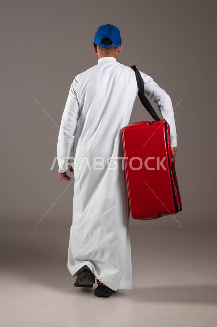 اداء العمل بدقة واتقان، التسوق وشراء الحاجيات عبر الانترنت، ضمان سلامة الطرود و البضائع المرسلة، بورتريه من الخلف لشاب عربي خليجي سعودي يرتدي الزي التقليدي  وقبعة زرقاء يحمل بيده حقيبة حمراء، عملية الشحن والتوصيل الموثوقه، صوره لكامل الجسم، خلفية رماديه