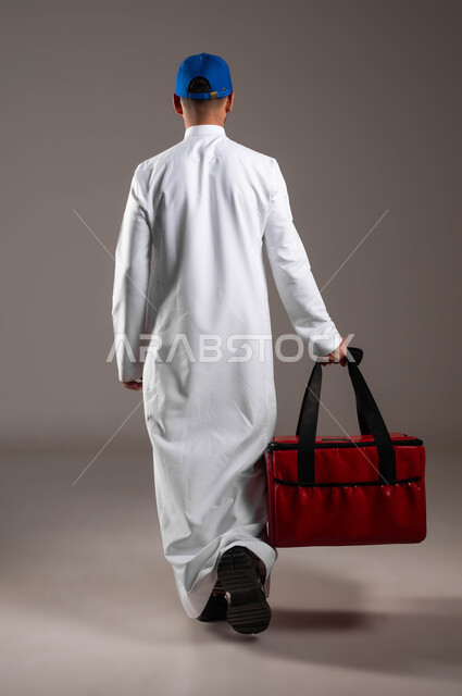 التسوق وشراء الحاجيات عبر الانترنت، ضمان سلامة الطرود و البضائع المرسلة، بورتريه من الخلف لشاب عربي خليجي سعودي يرتدي الزي التقليدي  وقبعة زرقاء يحمل بيده حقيبة حمراء، عملية الشحن والتوصيل الموثوقه، اداء العمل بدقة واتقان، صوره لكامل الجسم، خلفية رماديه