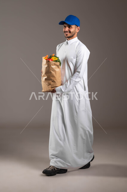 اداء العمل بدقة واتقان، منتجات وطنية محلية، ايماءات السعادة والسرور، شراء المواد الغذائية عبر الانترنت، خدمة التوصيل السريع في السعودية، بورتريه لمندوب عربي خليجي سعودي  يرتدي الزي التقليدي و قبعه زرقاء يحمل بيده كيس من الخضراوات، صورة لكامل الجسم، خلفيه رماديه