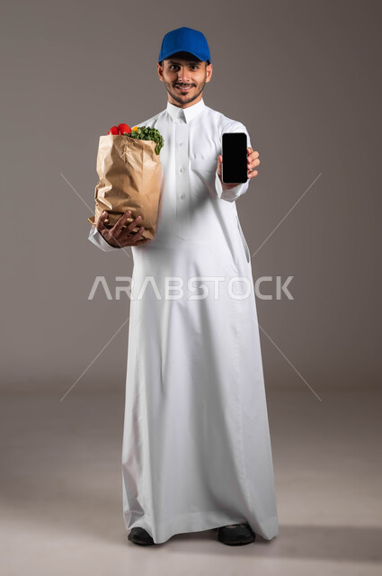 منتجات وطنية محلية، عرض شاشة الموبايل، الاستفادة من التكنولوجيا الحديثة بتسهيل العمل، خدمة التوصيل السريع في السعودية، بورتريه مقرب لمندوب عربي خليجي سعودي  يرتدي الزي التقليدي و قبعه زرقاء يحمل بيده كيس من الخضراوات، خلفيه رماديه