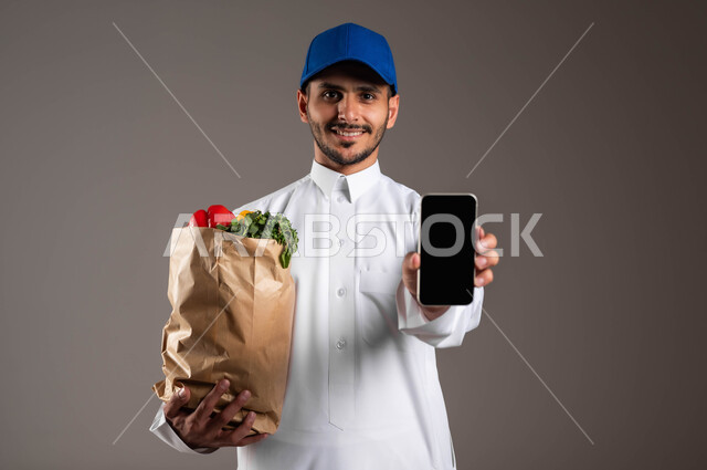 عرض شاشة الموبايل، الاستفادة من التكنولوجيا الحديثة بتسهيل العمل، منتجات وطنية محلية، خدمة التوصيل السريع في السعودية، بورتريه مقرب لمندوب عربي خليجي سعودي  يرتدي الزي التقليدي و قبعه زرقاء يحمل بيده كيس من الخضراوات، خلفيه رماديه