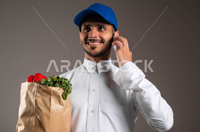 الاستفادة من التكنولوجيا الحديثة بتسهيل العمل، التواصل مع الزبائن عبر الموبايل، منتجات وطنية محلية، خدمة التوصيل السريع في السعودية، بورتريه مقرب لمندوب عربي خليجي سعودي  يرتدي الزي التقليدي و قبعه زرقاء يحمل بيده كيس من الخضراوات، خلفيه رماديه