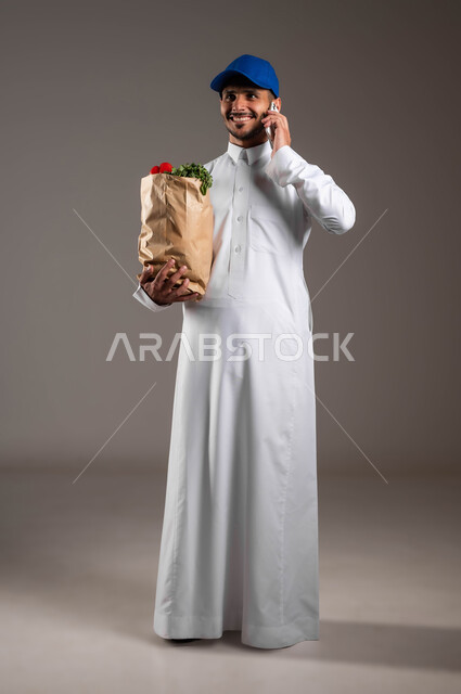 التواصل مع الزبائن عبر الموبايل، الاستفادة من التكنولوجيا الحديثة بتسهيل العمل، منتجات وطنية محلية، خدمة التوصيل السريع في السعودية، بورتريه لمندوب عربي خليجي سعودي  يرتدي الزي التقليدي و قبعه زرقاء يحمل بيده كيس من الخضراوات، صورة لكامل الجسم، خلفيه رماديه
