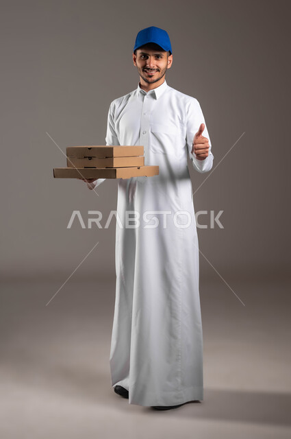 خدمة التوصيل السريعة والآمن في السعودية، بورتريه لمندوب عربي خليجي سعودي مبتسم يرتدي الزي التقليدي و القبعة الزرقاء يحمل بده مجموعة من الصناديق يرفع يده بايماءات الجودة، عملية نقل الطلبات للزبائن، صورة لكامل الجسم، خلفيه رماديه