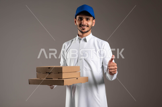 التسوق عن طريق الانترنت، خدمة التوصيل السريعة والآمن في السعودية، بورتريه مقرب لمندوب عربي خليجي سعودي مبتسم يرتدي الزي التقليدي و القبعة الزرقاء يحمل بده مجموعة من الصناديق يرفع يده بايماءات الجودة، عملية نقل الطلبات للزبائن، خلفيه رماديه