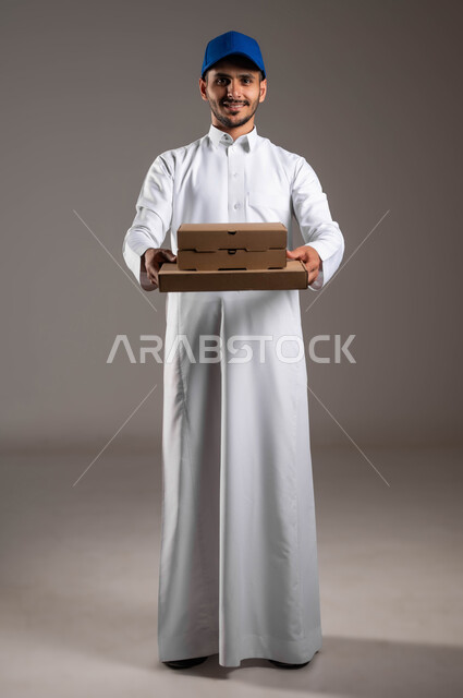 النظر إلى الكاميرا بإيماءات السعادة والسرور، بورتريه لمندوب توصيل عربي خليجي سعودي مبتسم يرتدي الزي التقليدي و القبعة الزرقاء يحمل بده مجموعة من الصناديق، خدمة التوصيل السريعة والآمن في السعودية، عملية نقل الطلبات للزبائن، صورة لكامل الجسم، خلفيه رماديه