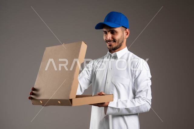 Buying necessities and shopping online, looking into the box with gestures of happiness and pleasure, the process of transferring orders to customers, the fast and safe delivery service in Saudi Arabia, a close-up portrait of a smiling Saudi Arabian Gulf representative wearing traditional clothing holding an open package in his hand, gray background.