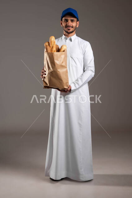 شراء الاحتياجات المنزلية والمواد الغذائية المختلفة من خلال الانترنت، المخبوزات والمعجنات الطازجة الشهية، خدمة التوصيل السريع في السعودية، بورتريه لمندوب عربي خليجي سعودي  يرتدي الزي التقليدي و قبعه زرقاء يقوم بتسلم كيس من الخبز، صورة لكامل الجسم، خلفيه رماديه